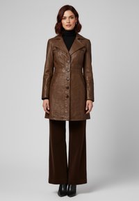 Bruine leren jas met grote kraag, zes knopen aan de voorkant en zijzakken; gecombineerd met wijde bruine broek en zwarte laarzen.