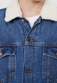 Detailní záběr na modrou denimovou bundu s bílým krkem z sherpy a kovovými knoflíky přes bílou košili.