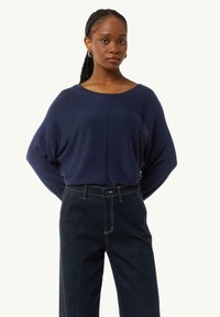 Navyblauer Strickpullover mit weitem Ausschnitt und lockerer Passform, kombiniert mit dunklen Jeans aus Denim, die über kontrastierende Nähte und einen Knopfverschluss verfügen.