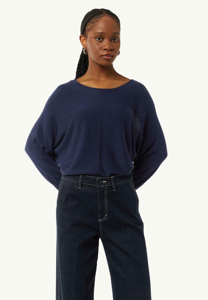 Navyblauer Strickpullover mit weitem Ausschnitt und lockerer Passform, kombiniert mit dunklen Jeans aus Denim, die über kontrastierende Nähte und einen Knopfverschluss verfügen.
