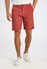 Shorts en coton rouges avec des revers, une fermeture à bouton sur le devant et des poches latérales. Portés avec des baskets blanches sur un fond neutre.
