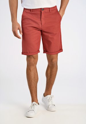 Rote Baumwollshorts mit umgeschlagenen Säumen, einem vorderen Knopfverschluss und seitlichen Taschen. Getragen mit weißen Sneakers vor einem neutralen Hintergrund.