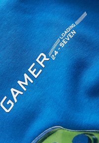 T-shirt en coton bleu avec le texte blanc "GAMER" et "CHARGEMENT 24-HEURES". Le design comprend un élément graphique vert en bas.