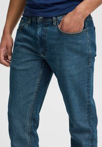 Jeans de denim azul con corte recto, que cuentan con cinco bolsillos, detalles de costura sutiles y una textura ligeramente desgastada.