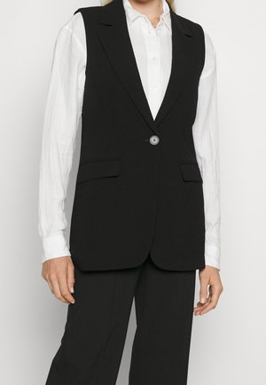 Frau trägt einen schwarzen, maßgeschneiderten ärmellosen Blazer über einem weißen, langärmeligen Hemd und schwarzen Hosen, steht vor einem schlichten Hintergrund.