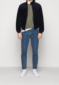 Black velour jacket with high collar, red logo, green crew neck sweater, blue jeans, and white sneakers. Smooth texture and fitted design.