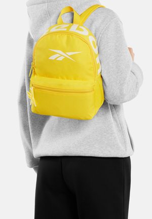 Osoba nosząca jaskrawożółty plecak Reebok z białymi logo, na jasnoszarej bluzie z kapturem i czarnych spodniach, widziana od tyłu.