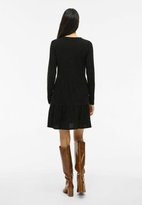 Robe noire à côtes avec des manches longues, haut ajusté et jupe évasée. Assortie à de grandes bottes marron brillantes qui ajoutent de la texture et du contraste.