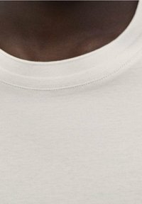 Närbild av en person med mörk hudton som bär en enkel ljusbeige rundhalsad t-shirt, med fokus på hals- och övre bröstområde.