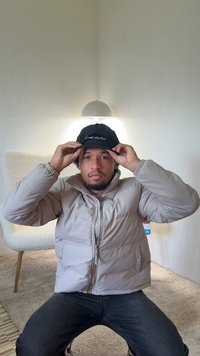 Svart lue med hvit tekst, lys beige dunjakke, mørke jeans og nøytral bakgrunn med myk belysning. Fokus på teksturer og farger.