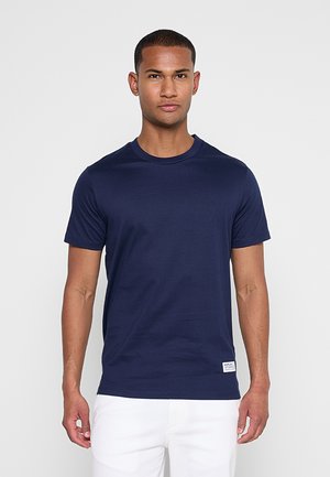 Mann trägt ein schlichtes dunkelblaues T-Shirt mit kurzen Ärmeln und weiße Hosen, steht vor einem hellgrauen Hintergrund.