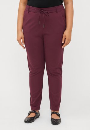 Kvinde iført bordeaux slim-fit bukser med snøre i taljen og sorte Mary Jane-sko med nitter, stående mod en hvid baggrund.