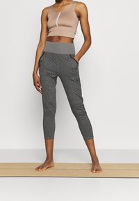Mujer de pie descalza sobre una esterilla de yoga, vestida con un top corto beige y mallas deportivas grises de cintura alta con bolsillos.
