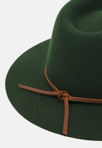 Zöld filc kalap széles karimával és lekerekített tetejével. Egy csomózott barna bőrszíj díszíti az alját. Puha tapintású és sima felületű.