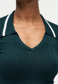 Gros plan d'une personne portant un pull en maille torsadée vert foncé avec un col en V et un col orné de rayures blanches.