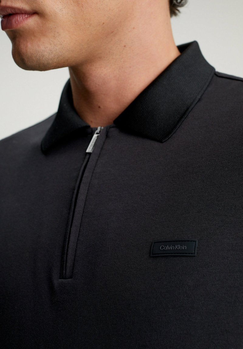 Donkergrijze polo met een ritssluiting bij de kraag, geribbelde kraagaccenten en een klein zwart label met "Calvin Klein" aan de voorkant.