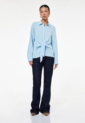 Kobieta stojąca, ubrana w jasnoniebieską, długorękawową koszulę z guzikami i wiązaniem z przodu, ciemne dzwonowe jeansy, brązowe spiczaste buty oraz złote kolczyki koła.