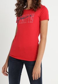 Mujer con una camiseta roja ajustada que tiene el logotipo "PEPE" en texto blanco y azul marino contorneado, combinada con jeans azul oscuro, contra un fondo blanco.