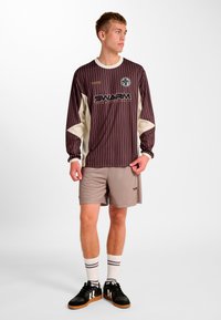 Braunes langärmliges gestreiftes Hemd mit weißen Akzenten, Logo und Text, kombiniert mit grauen Shorts, schwarzen Turnschuhen und weißen gestreiften Socken.