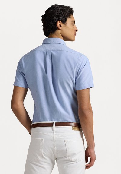 Chemise légère à manches courtes de couleur bleu clair avec un col boutonné, en tissu texturé, et une pince au dos, associée à un pantalon blanc et une ceinture en cuir marron.