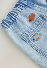 Material din denim albastru deschis, cu talie elastică și doi pești colorați brodați lângă buzunar.