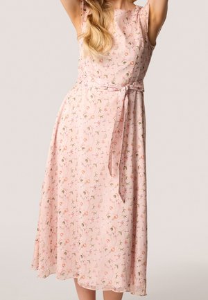 Femme portant une robe midi rose fleurie sans manches avec une ceinture à la taille, debout devant un fond clair uni avec les bras levés.