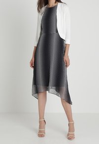 Vestido a lunares blanco y negro con un dobladillo asimétrico, combinado con un cárdigan corto blanco. Tacones nude con tiras completan el look.