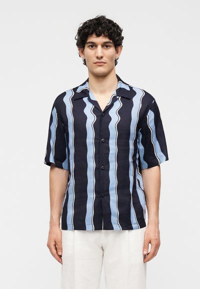 Jeune homme aux cheveux courts et bouclés portant une chemise à manches courtes boutonnée à rayures ondulées bleu marine et bleu clair et un pantalon blanc.