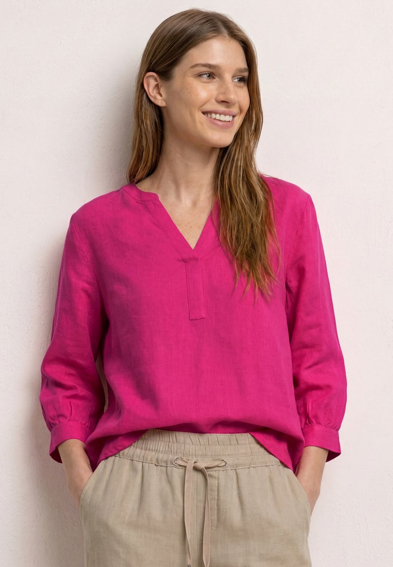 Jeune femme souriante, portant un chemisier rose vif à manches longues et un pantalon beige avec cordon, debout devant un mur clair texturé.