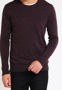 Burgunderfarbener Pullover aus weichem Material, mit Rundhalsausschnitt, langen Ärmeln und einer Brusttasche. Gerippte Bündchen und Saum.