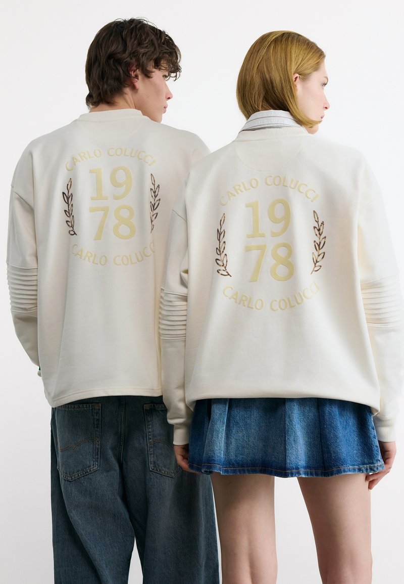 Δύο άτομα γυρισμένα από την πλάτη, φορούν ίδια λευκά φούτερ με το μήνυμα «CARLO COLUCCI 1978» και σχέδια δάφνης στην πλάτη, συνδυασμένα με τζιν παντελόνια και τζιν φούστα.