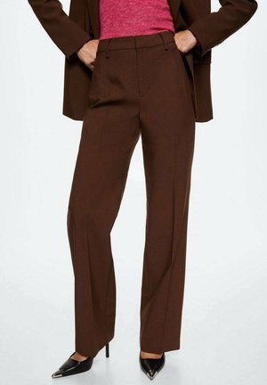 Persoon die hoge, brede bruine broek draagt, zwarte puntige hakken, een roze top en een bijpassend bruine blazer met handen in de zakken.