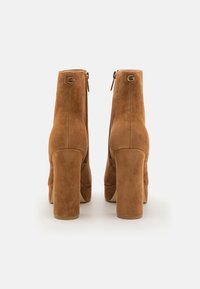 Stivaletti alla caviglia con tacco a blocco in suede color nocciola, con cerniere sul retro, dettagli in logo dorato e una texture liscia.