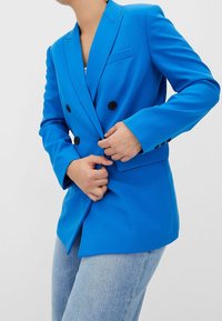 Blazer bleu croisé en tissu lisse, doté de boutons noirs, d'un revers à cran et de poches plaquées latérales. Porté avec du denim.