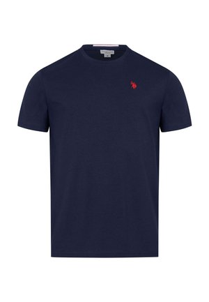 Mann mit dunklen Haaren und Bart steht mit verschränkten Armen und trägt ein dunkelblaues T-Shirt mit kleinem rotem Logo auf der Brust.