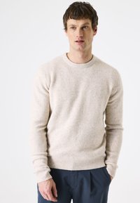 Licht beige gebreide trui van zacht materiaal, met een ronde halslijn en geribbelde boorden en zoom, gedragen met donkerblauwe broek.