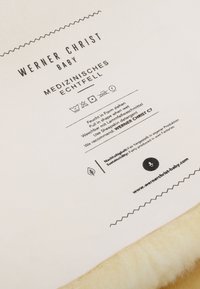 Medizinisches Lammfell-Produktetikett mit schwarzem Text auf hellem Hintergrund, das Pflegeanweisungen, Informationen zur Nachhaltigkeit und den Markennamen enthält.