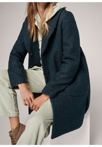 Tumma petroolinsininen teksturoitu villakangastakki rentolla istuvuudella, isoilla kauluksilla ja kahdella napilla. Päällä vaaleanvihreä huppari ja corduroyhousut.