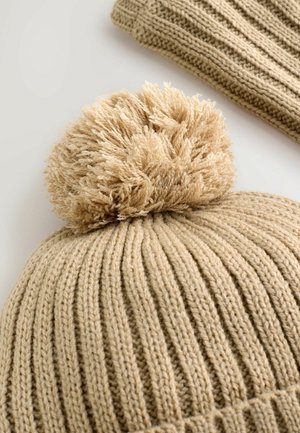 Beige kötött sapka bordás mintával, tetején egy bolyhos pom-pommal. Puha textúrával és meleg anyaggal rendelkezik, ami alkalmas a hideg időjárásra.