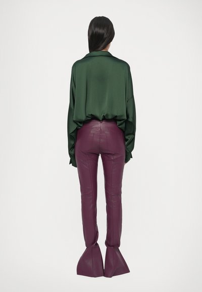 Zielona, satynowa koszula oversize z kołnierzykiem w zestawieniu z dopasowanymi, fioletowymi spodniami z poliuretanu o rozszerzanych nogawkach. Gładka faktura i nowoczesne detale.