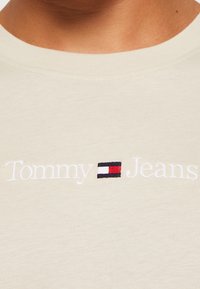 Béžové bavlněné tričko s kulatým výstřihem, na kterém je bílé vyšívané textové logo "Tommy" a vedle něj malé logo v námořnické modré, červené a bílé barvě.