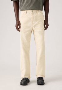 Μπεζ παντελόνι σε ίσια γραμμή με λεία υφή, διαθέτει κούμπωμα  με κουμπί και τσέπες. Φοριέται με ριγέ κοντομάνικη μπλούζα και μαύρα αθλητικά παπούτσια.