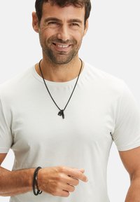 Zwarte ketting met twee geometrische hangers, gedragen met een eenvoudig wit T-shirt; gevlochten zwarte armband om de pols.