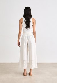 Pantalons cropped en coton blanc avec un design à jambes larges, ornés de broderies et de détails en dentelle au niveau de l'ourlet. Associés à un débardeur blanc ajusté.