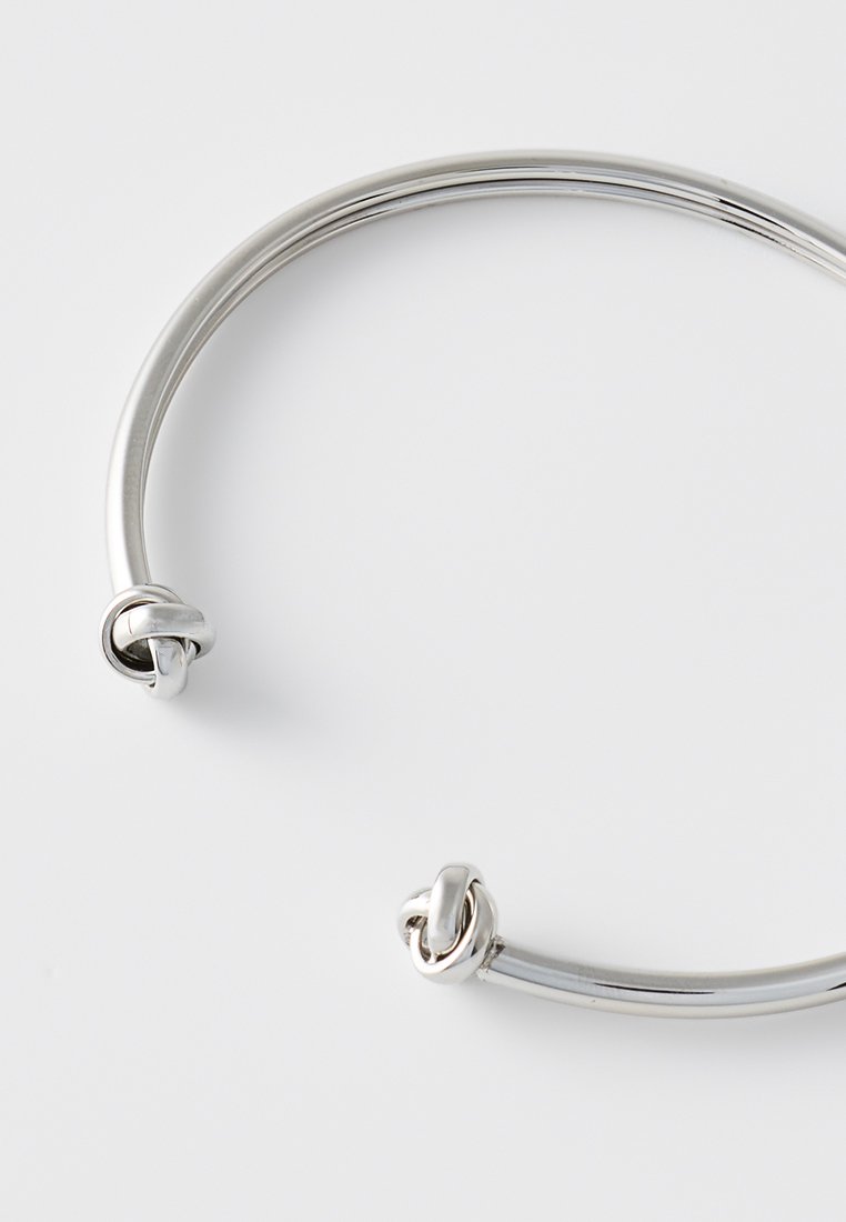 Bracciale in metallo argento con un design liscio e arrotondato, caratterizzato da due nodi intrecciati a ciascun estremo. Finitura lucida con un aspetto elegante.