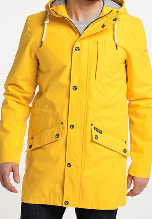 Lysegul hættejakke med trykknapper, brystlomme og to store lommer med klapper og knapper båret af en person.