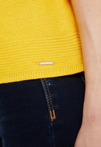 Gelb gestrickter Pullover mit Rippstruktur und einem kleinen silbernen Logotag am Saum, kombiniert mit dunklen Denim-Jeans mit kontrastierenden Nähten.