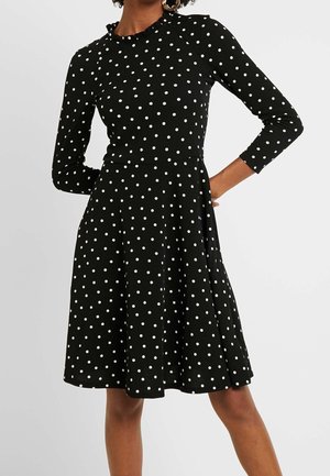 Robe noire à pois blancs, manches longues, avec un col à volants. Jupe évasée arrivant au niveau des genoux, faite en tissu lisse.