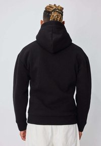 Personne aux cheveux tressés attachés en arrière, portant un sweat à capuche noir et un pantalon blanc, tournée de dos sur un fond uni gris clair.