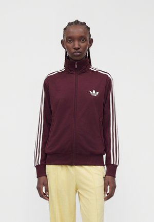 Osoba s vrkočmi, ktorá nosí bordovú tréningovú bundu Adidas s bielymi pruhmi a logami, v kombinácii so svetložltými nohavicami, stojí na jednoliatom pozadí.